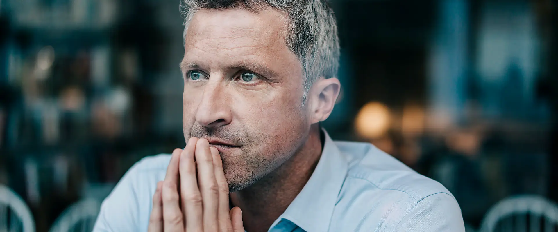Nachdenklicher Mann mit grauen Haaren und blauen Augen, der die Hände vor dem Gesicht faltet. Er trägt ein hellblaues Hemd und sitzt in einem stilvollen, modernen Raum.