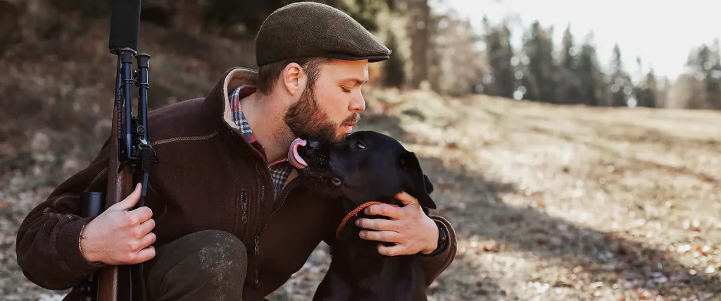 Junger Mann mit Schusswaffe und Jagdhut küsst seinen schwarzen Hund in der Natur.