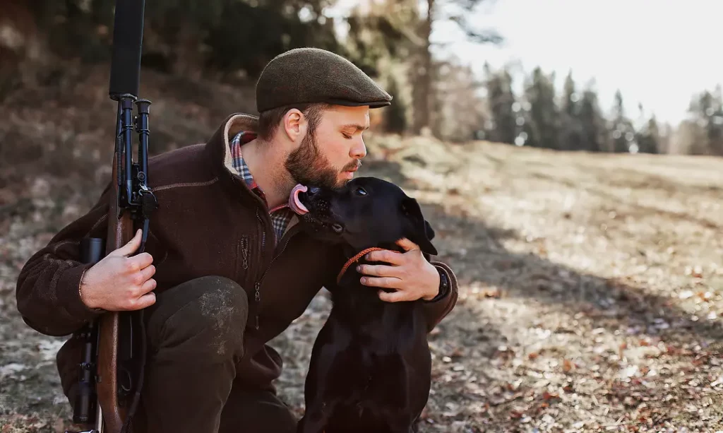 Mann mit Schusswaffe umarmt und küsst seinen Hund in einer natürlichen Umgebung.