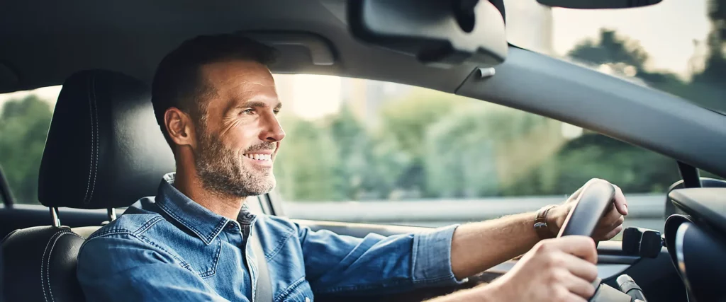 Lächelnder Mann fährt ein Auto, trägt ein Jeanshemd und hat beide Hände am Lenkrad.