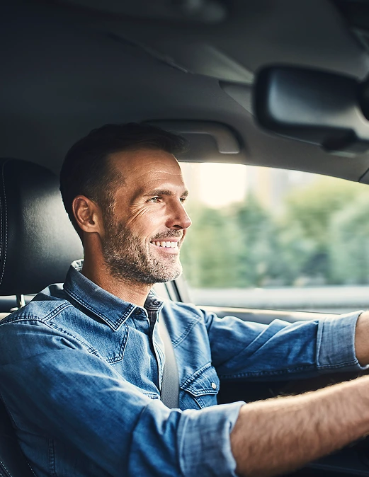 Lächelnder Mann fährt ein Auto, trägt ein Jeanshemd und hat beide Hände am Lenkrad.